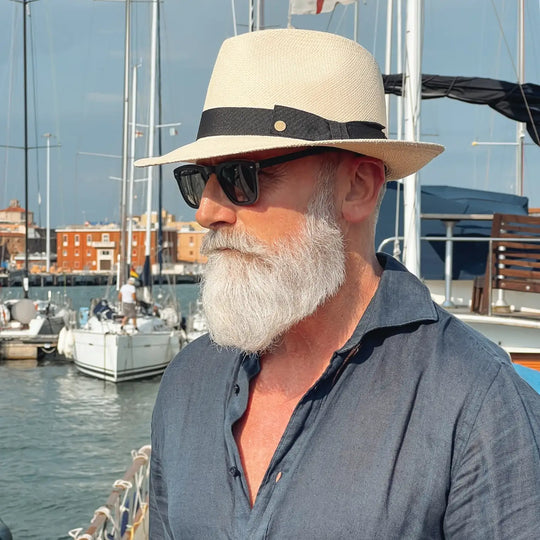 vista a tre quarti di un signore adulto che ricorda un gentleman di altri tempi, al porto, con barba lunga bianca, occhiali da sole, camicia blu di lino e pantaloni bianchi, che indossa il cappello di panama con vista frontale in stile fedora color naturale di Primario Nesti