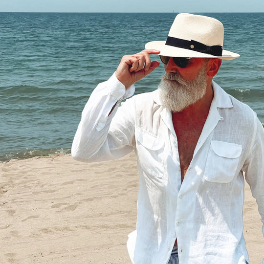 vista a tre quarti di un signore adulto che ricorda un gentleman di altri tempi, al mare, con barba lunga bianca, occhiali da sole e camicia bianca di lino che indossa il cappello di panama con vista laterale a sinistra  in stile fedora color naturale di Primario Nesti