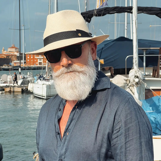 vista a tre quarti di un signore adulto che ricorda un gentleman di altri tempi, al porto, con barba lunga bianca, occhiali da sole, camicia blu di lino un po’ sbottonata e pantaloni bianchi, che indossa il cappello di panama con vista frontale in stile fedora color naturale di Primario Nesti