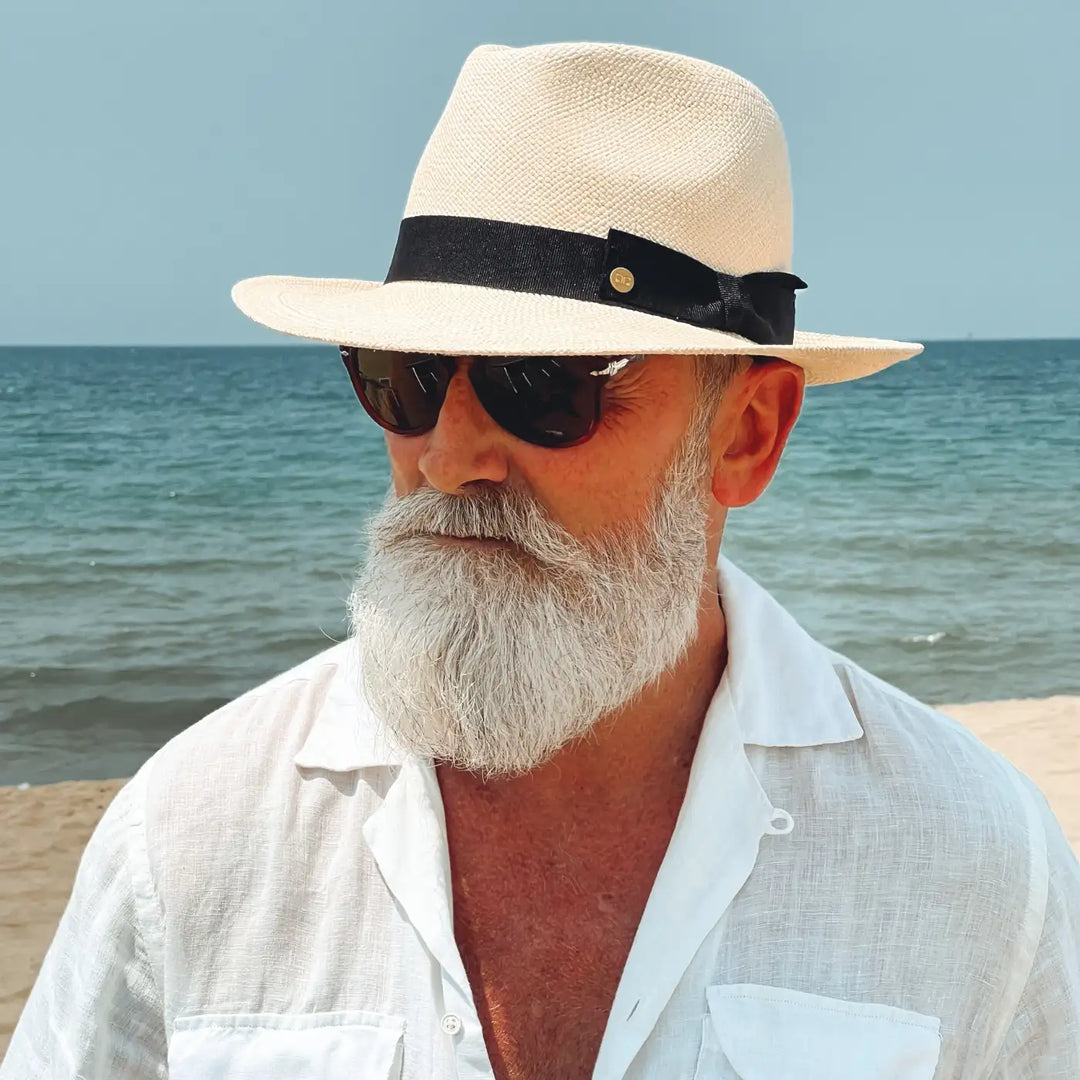 primo piano di un signore adulto che ricorda un gentleman di altri tempi, al mare, con barba lunga bianca, occhiali da sole e camicia bianca di lino che indossa il cappello di panama in stile fedora color naturale di Primario Nesti