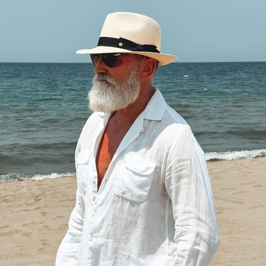 mezzo busto spostato verso sinistra di un signore adulto che ricorda un gentleman di altri tempi, al mare, con barba lunga bianca, occhiali da sole e camicia bianca di lino che indossa il cappello di panama in stile fedora color naturale di Primario Nesti