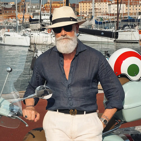 primo piano laterale a sinistra di un signore adulto che ricorda un gentleman di altri tempi, al porto, appoggiato a una vespa, con barba lunga bianca, occhiali da sole, camicia blu di lino e pantaloni bianchi, che indossa il cappello di panama in stile fedora color naturale di Primario Nesti