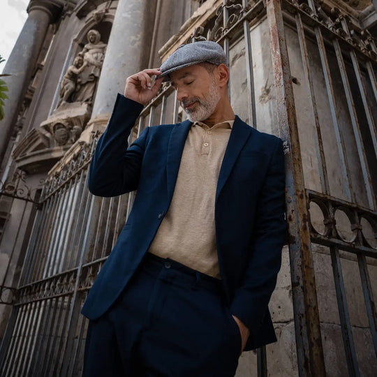 modello adulto con barba indossa una coppola peaky blinder spigata primario nesti in un look navy e beige con cancello storico