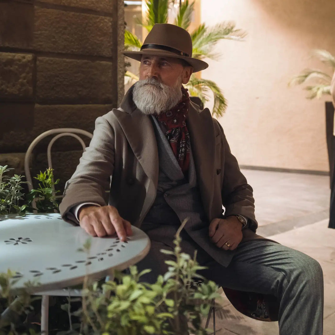 modello distinto con cappello fedora classico primario nesti antipioggia colore deserto tenue e cappotto a tono in location outdoor con elementi naturali
