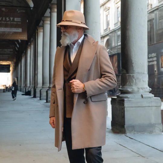 modello elegante con cappello fedora tesa corta primario nesti antipioggia colore beige indossa un completo beige scuro con sfondo di colonne storiche