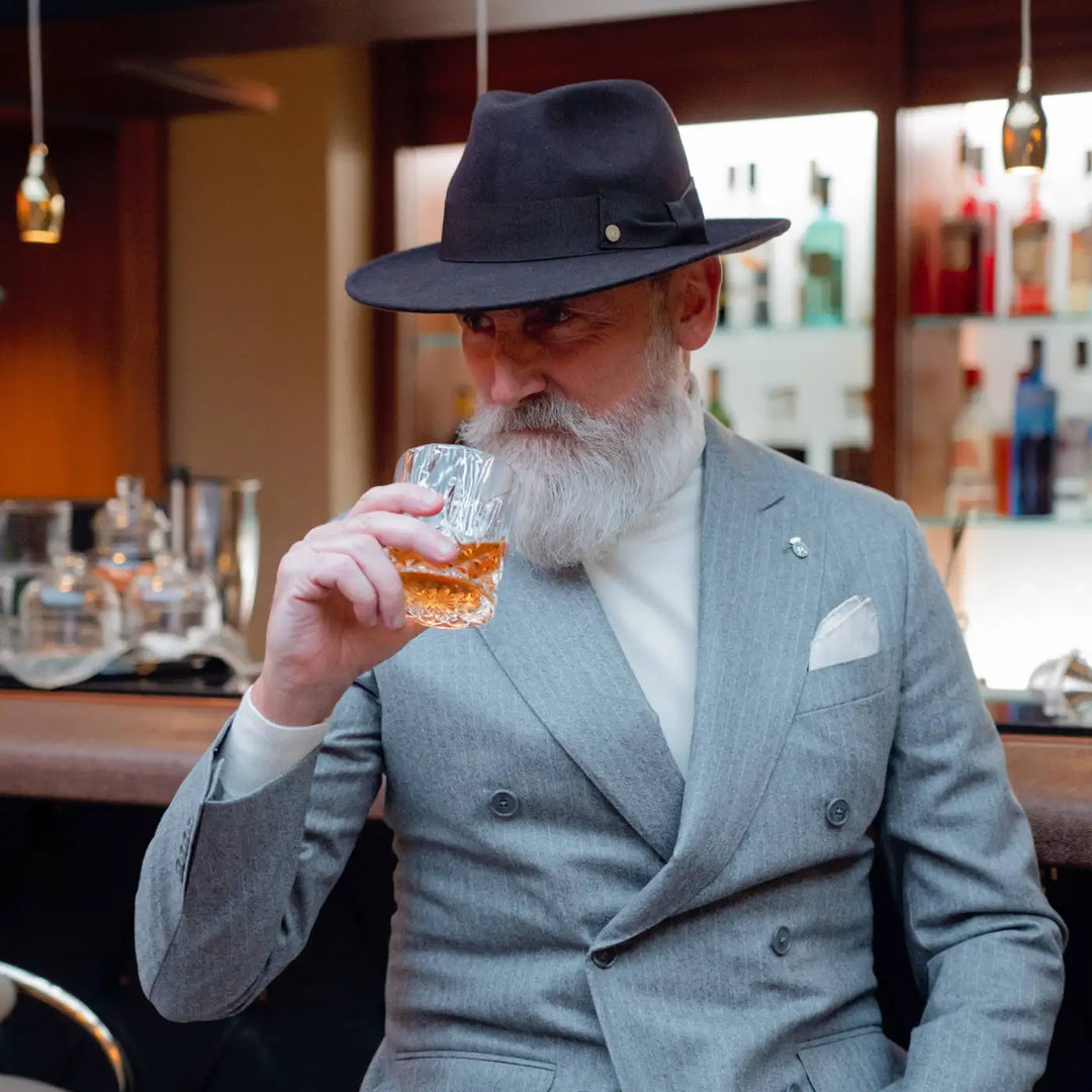 modello elegante con fedora coccos primario nesti in lana merinos colore antracite e completo grigio chiaro assora del whisky in un bar sofisticato