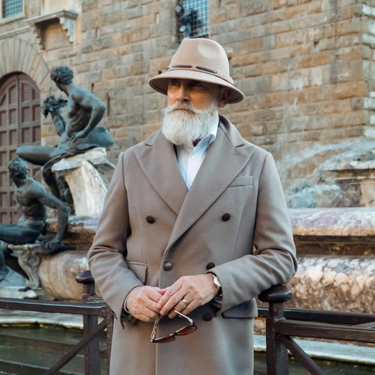 modello maturo con cappello fedora tesa corta primario nesti antipioggia colore beige indossa un completo beige scuro per uno stile italiano raffinato tra colonne storiche