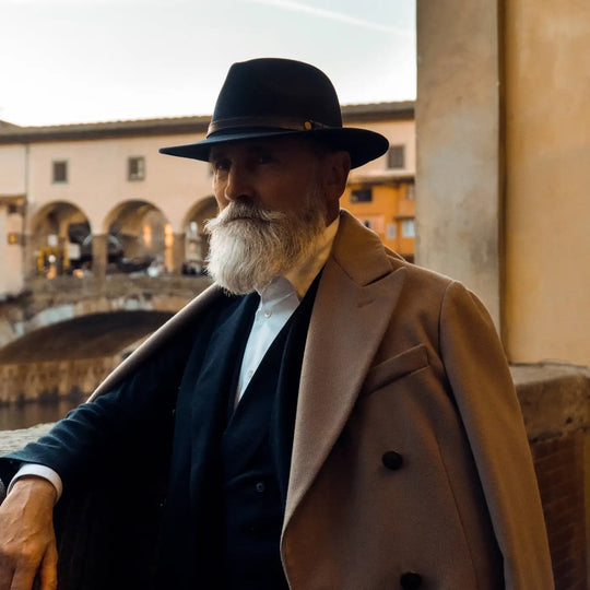 modello maturo con cappello fedora tradizionale primario nesti antipioggia colore blu con vista ponte vecchio indossa un completo scuro elegante per look italiano sofisticato