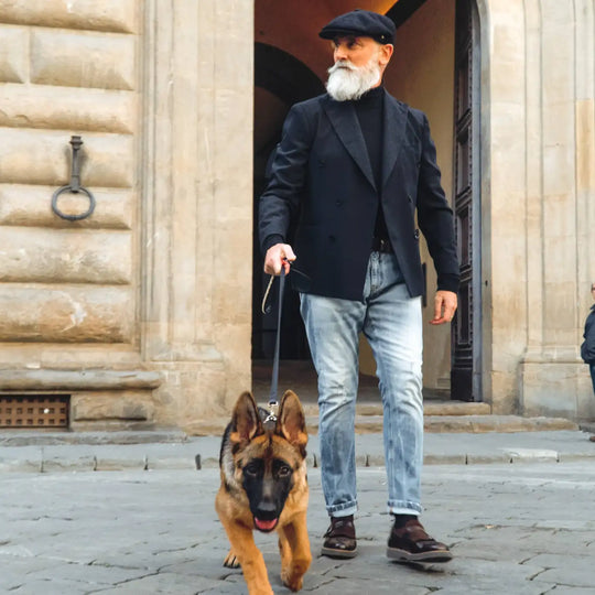 modello maturo con coppola peaky blinders primario nesti blu in un look casual con jeans e cane pastore tedesco street style toscano autentico davanti palazzo storico