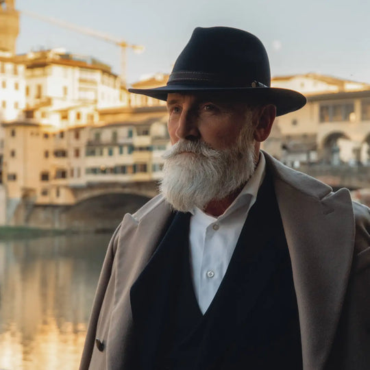 modello maturo raffinato con cappello fedora tradizionale primario nesti antipioggia indossa un completo nero e cappotto beige con ponte vecchio sullo sfondo