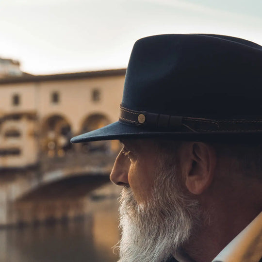 profilo elegante di modello con barba curata e cappello fedora tradizionale primario nesti antipioggia blu immerso nei riflessi dorati del tramonto sullarno